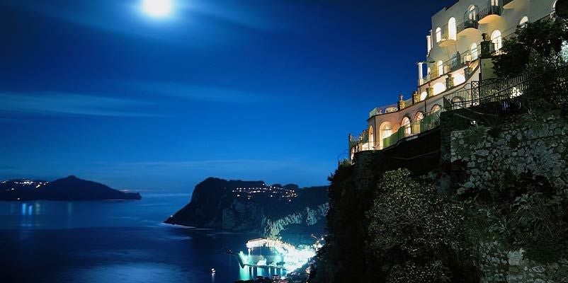 Luxury cliffside hotel overlooking Bay of Naples, with elegant terraced gardens, infinity pool, and panoramic views of Capri island at sunset