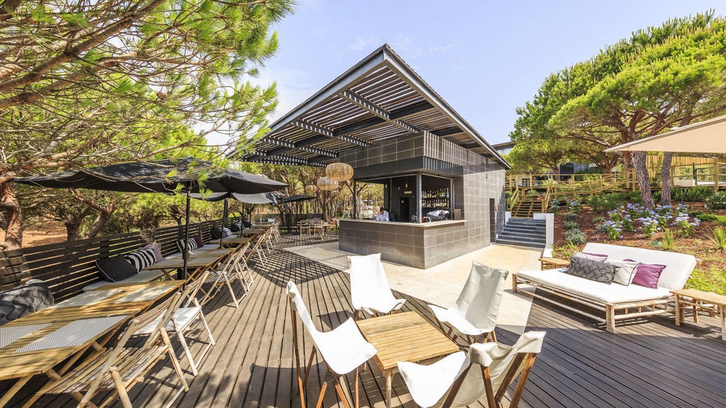 Modern beachfront hotel with sleek white architecture, surrounded by lush pine forest and overlooking pristine sandy beach at sunset