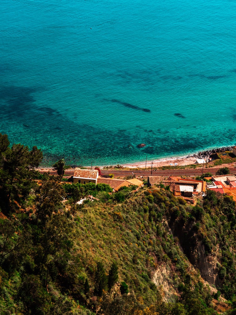 Mediterranean coastal panorama of Taormina, Sicily, featuring ancient stone buildings perched on cliffs above turquoise waters at sunset