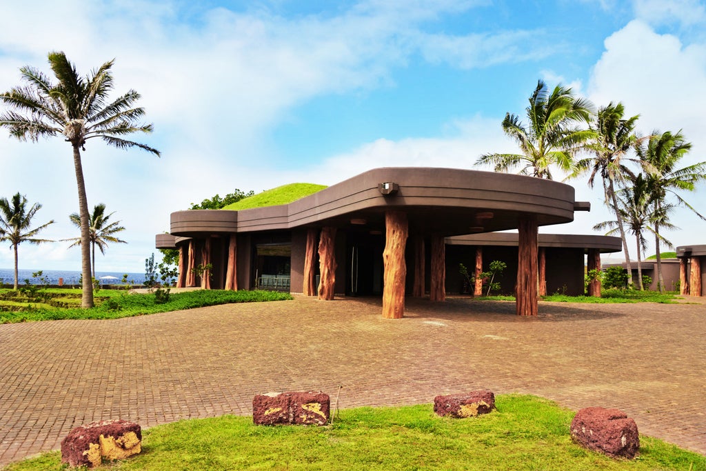 Curved, modern eco-resort with thatched roofs nestled into green hillside overlooking Pacific Ocean at Easter Island's luxury Hangaroa