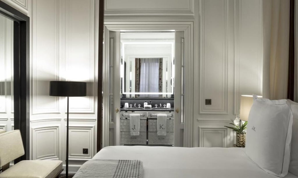 Elegant Parisian hotel room with plush ivory bedding, ornate gold-framed mirror, soft neutral palette, luxury furnishings and refined architectural details