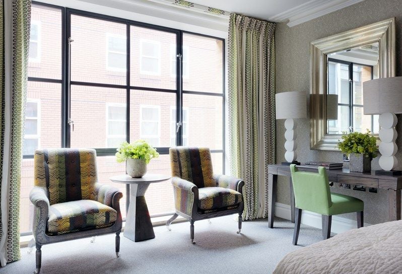 Elegant light-filled luxury room at Ham Yard Hotel featuring plush seating, vibrant textiles, contemporary artwork, and floor-to-ceiling windows with city view.