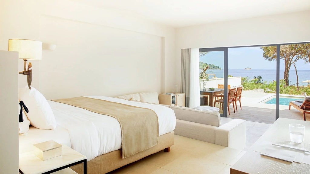 Luxurious beachfront junior suite at ELIVI Skiathos, featuring elegant white decor, plush bed, and panoramic sea view through expansive glass doors in Greece.