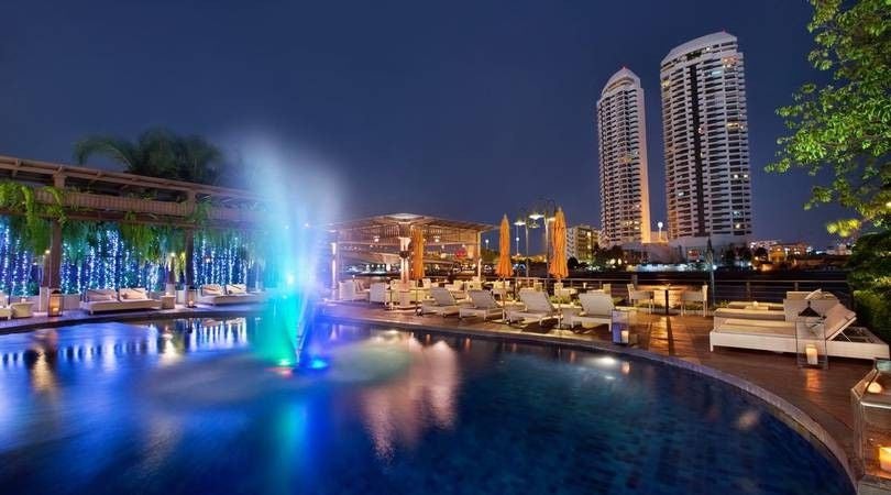 Elegant riverside boutique hotel in Bangkok with infinity pool overlooking Chao Phraya river, surrounded by tropical plants at sunset