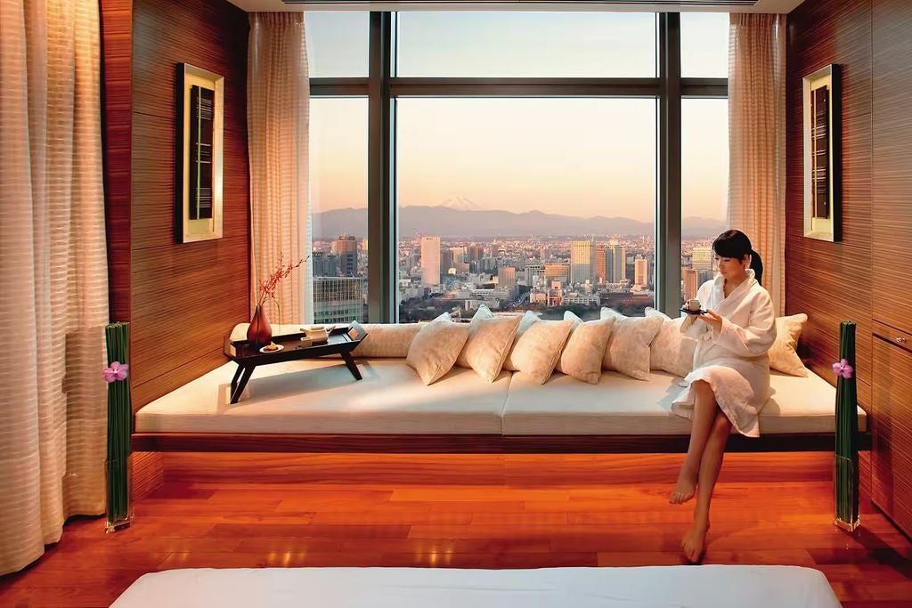 Modern luxury high-rise hotel tower with floor-to-ceiling windows overlooking Tokyo's skyline at dusk, illuminated by warm golden lights