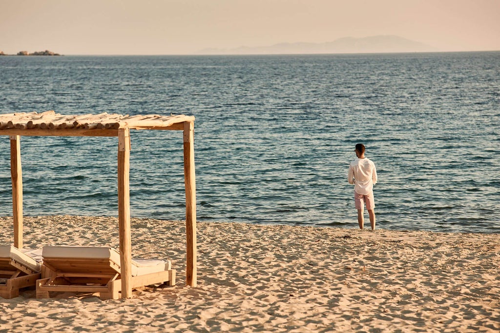 Luxurious white-washed beachfront hotel with minimalist Cycladic architecture, azure Aegean Sea, and pristine sandy shoreline at sunset