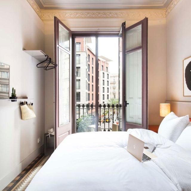 Minimalist Barcelona hotel room with crisp white linens, modern wooden furniture, large window, and soft natural light illuminating elegant interior design