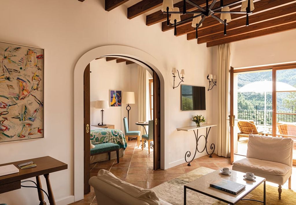 Sunlit luxury suite with rustic stone walls, elegant seating area, exposed wood beams, and panoramic windows overlooking Mallorca mountains