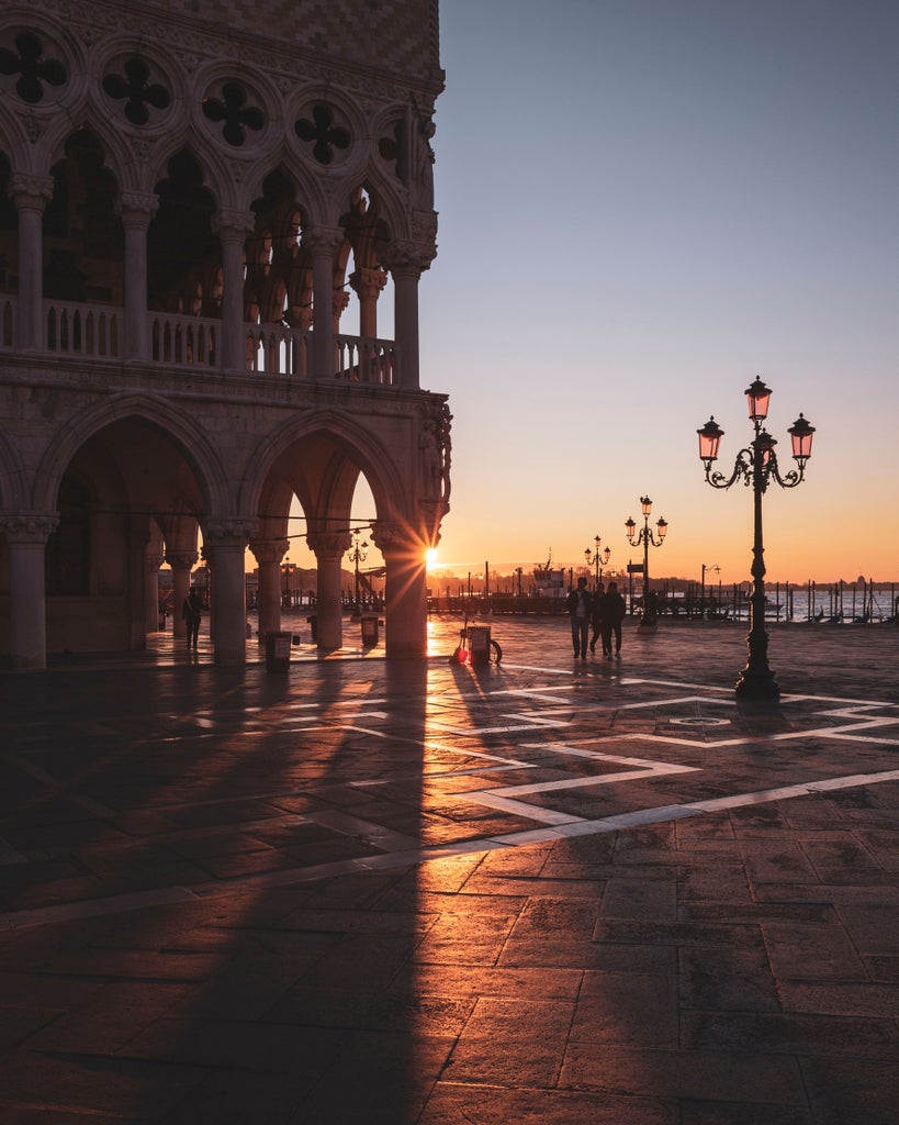 Traditional wooden gondola glides through narrow Venice canal at sunset, flanked by historic pastel buildings and ornate bridges