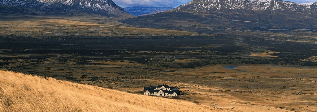 Luxurious stone lodge EOLO nestled in Patagonian steppe, with panoramic windows facing snow-capped mountains and golden grasslands