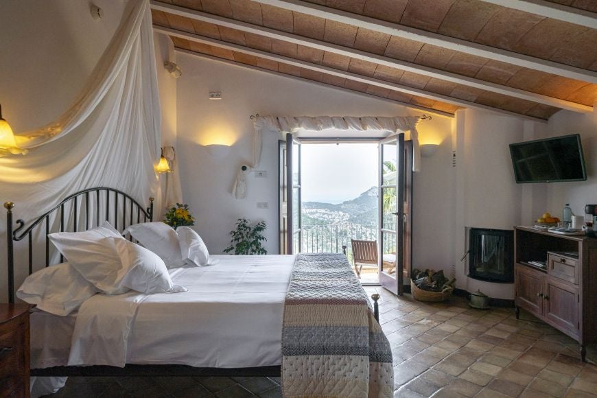 Elegant stone-walled deluxe bedroom with rustic wooden furniture, soft white linens, and panoramic view of Spanish countryside landscape