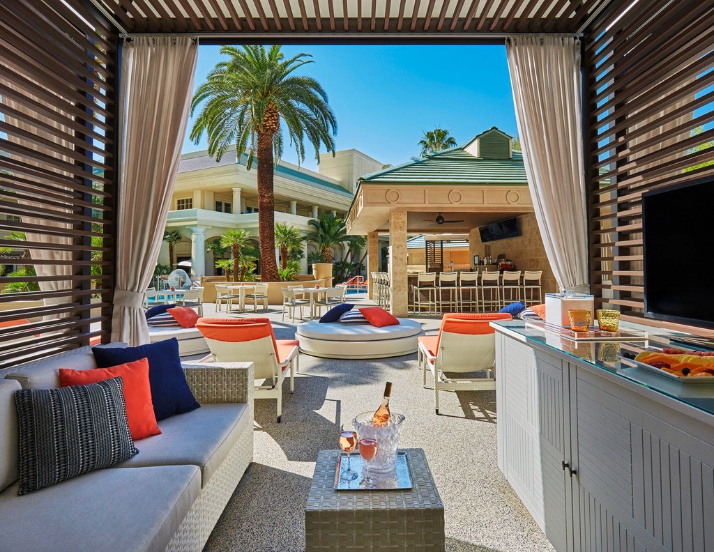 Modern luxury hotel tower with glass facade, twin fountains at entrance, palm trees and desert landscaping under blue Las Vegas sky