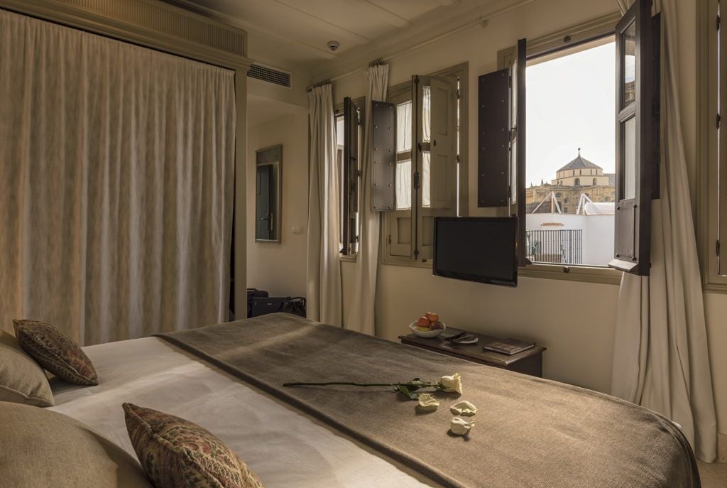 Elegant Spanish colonial-style hotel room with antique wooden furniture, ornate white walls, and large window overlooking historic Córdoba cityscape