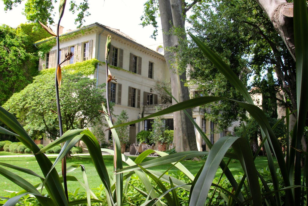 Elegant French boutique hotel facade with ornate wrought-iron balconies, soft pastel exterior, and lush garden with blooming flowers against blue sky