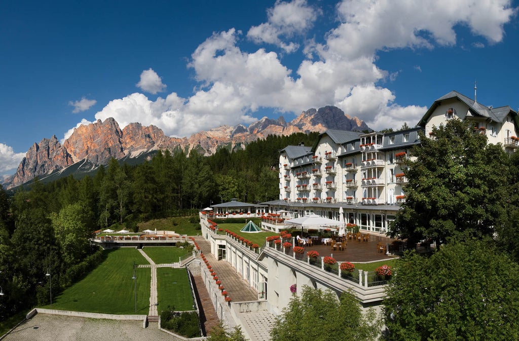 Elegant mountain resort with curved white facade, grand windows, and snow-capped Dolomites backdrop against clear blue sky