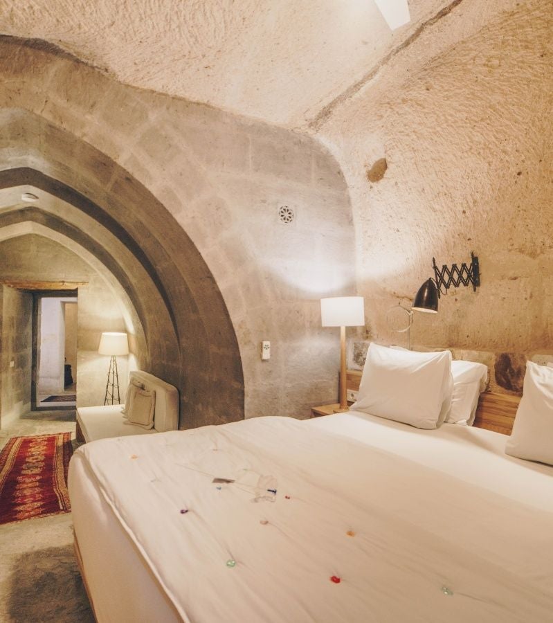 Elegant stone-walled suite with plush white bedding, rustic wooden furniture, and arched windows overlooking Cappadocia's unique volcanic landscape at sunset