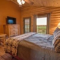 Rustic wooden cabin bedroom with plush queen bed, large windows overlooking scenic mountain landscape, warm earth-toned decor, and natural wood furnishings