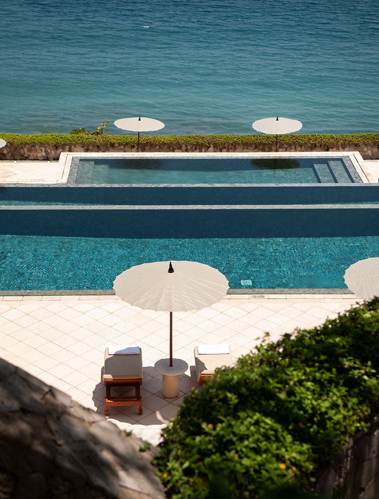 Three-tiered infinity pools cascade down a lush hillside overlooking the Bali Sea, with traditional thatched-roof pavilions nestled among palm trees