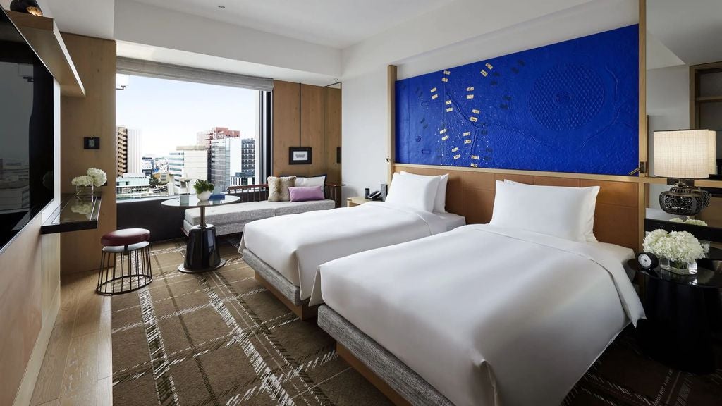 Clean, modern hotel room with two plush twin beds, floor-to-ceiling windows offering city views, contemporary art, and minimalist decor