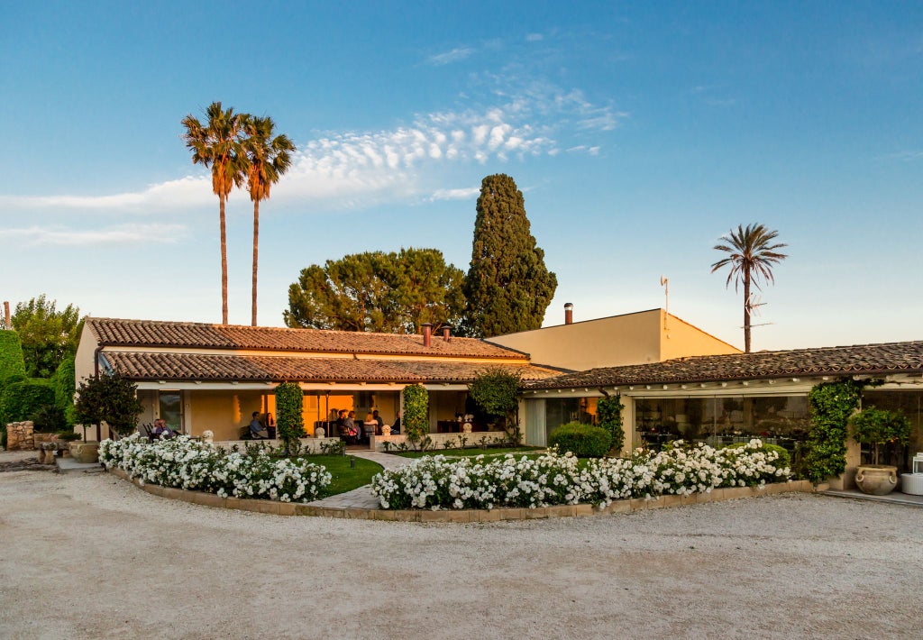 Elegant Sicilian luxury suite with soft neutral tones, ornate headboard, crisp white linens, and traditional architectural details overlooking lush garden landscape