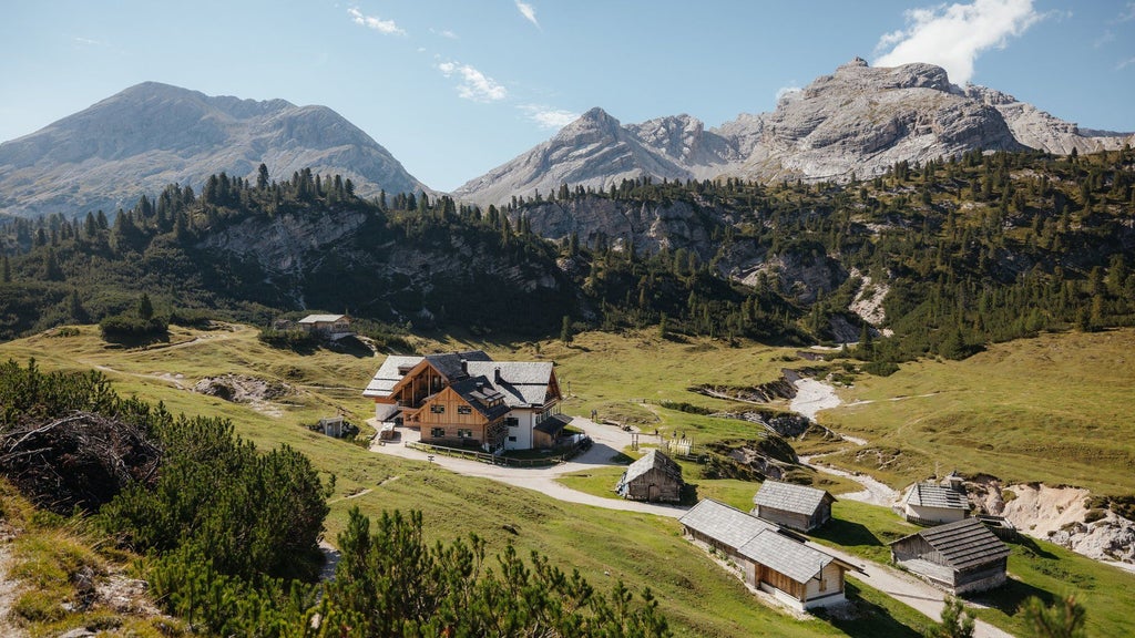 Traditional alpine wooden mountain lodge with wrap-around balconies nestled in lush green meadows beneath dramatic Dolomite peaks