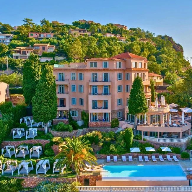 Luxurious Mediterranean coastal hotel with white exterior, blue-green sea backdrop, elegant balconies, and lush tropical landscaping in French Riviera setting