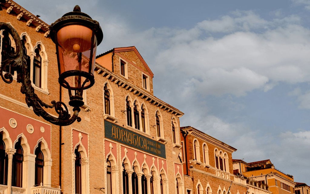 Elegant Italian palazzo hotel exterior with limestone facade, arched windows, and lush terracotta-tiled rooftops overlooking a serene Venetian canal at sunset