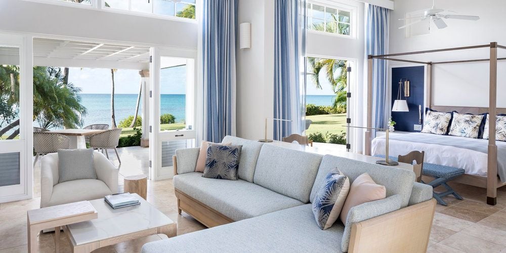 Luxurious beachside suite with pristine white linens, wooden accents, and panoramic views of turquoise Caribbean waters at Jumby Bay Island Resort in Antigua