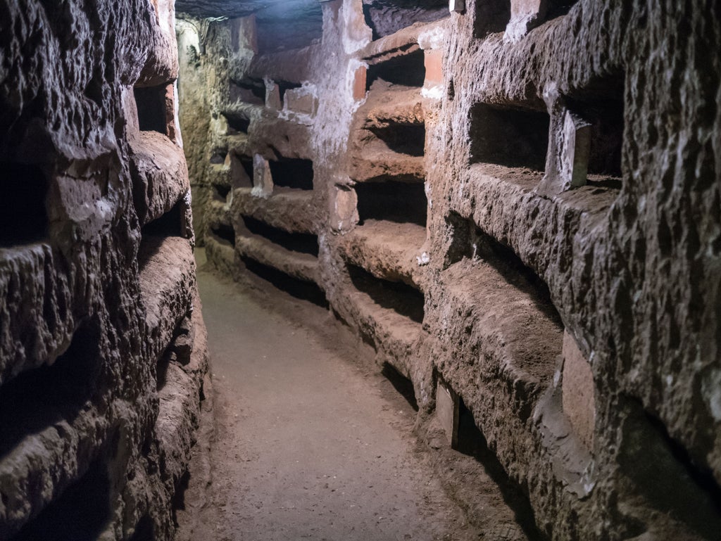 Cyclists explore ancient cobblestones of Appian Way amid towering umbrella pines, leading to ornate catacomb entrance in Rome