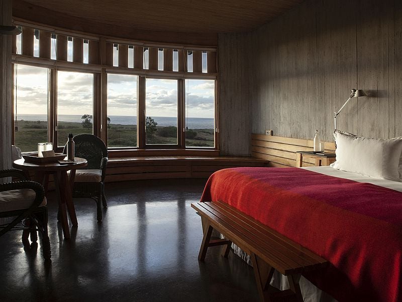 Minimalist luxe suite with panoramic ocean views, featuring sleek wooden design, white linens, and floor-to-ceiling windows overlooking Easter Island's rugged landscape