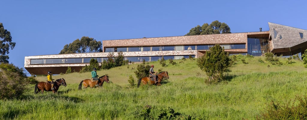 Modern wooden eco-lodge perched on coastal cliffs, featuring floor-to-ceiling windows and panoramic views of Chiloé archipelago at sunset