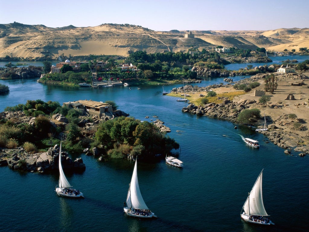 Traditional wooden dahabiya boat on Nile River with elegant cream-colored sails, ornate brass details, and teak wood deck at golden hour