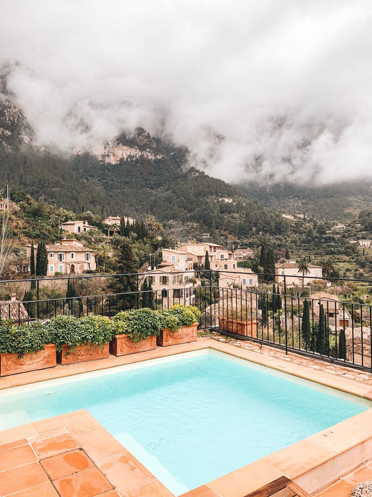 Mediterranean luxury hotel nestled in mountains, featuring stone buildings, lush gardens, and a tranquil swimming pool with mountain views