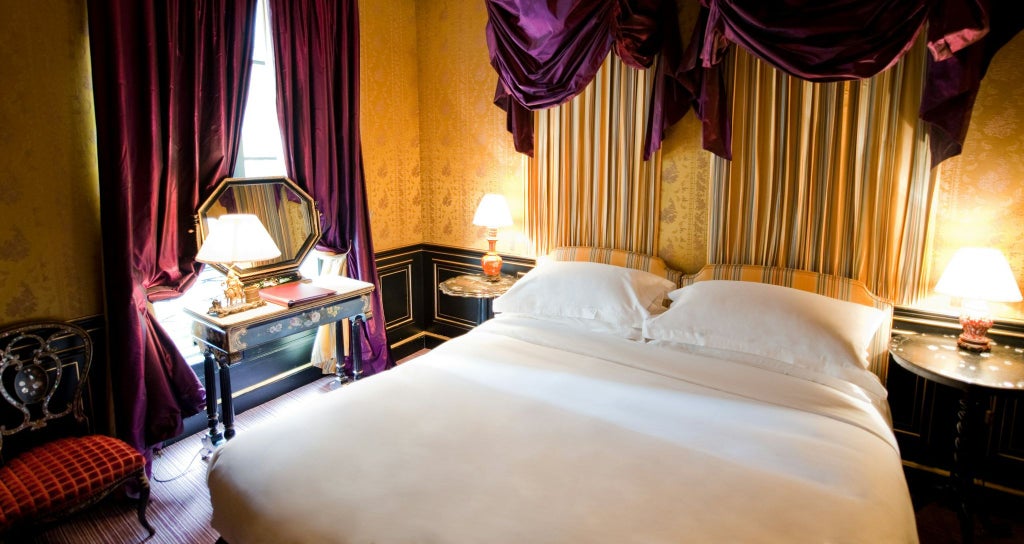 Elegant French boutique hotel room with plush king bed, ornate crystal chandelier, and rich burgundy accent wall overlooking Paris cityscape