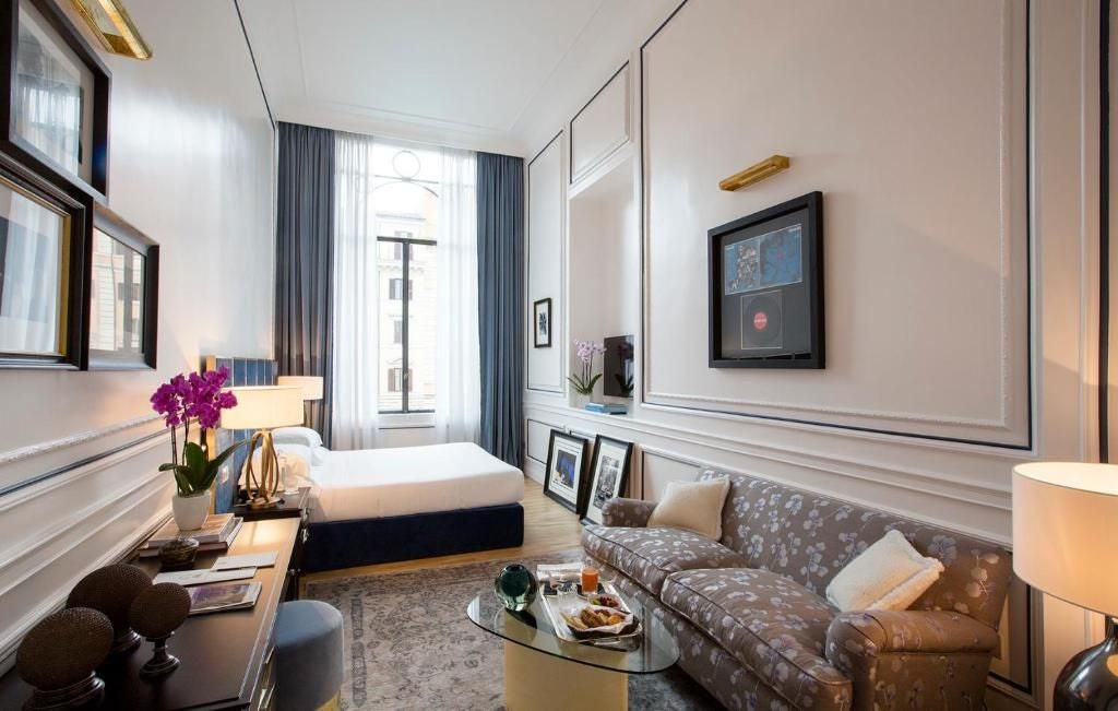 Elegant Italian luxury hotel room with plush white bed, marble details, modern artwork, and large windows overlooking scenic urban landscape in Rome