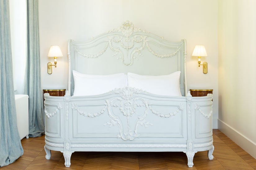 Elegant French château hotel room with king bed, crystal chandelier, ornate moldings and floor-to-ceiling windows overlooking manicured gardens