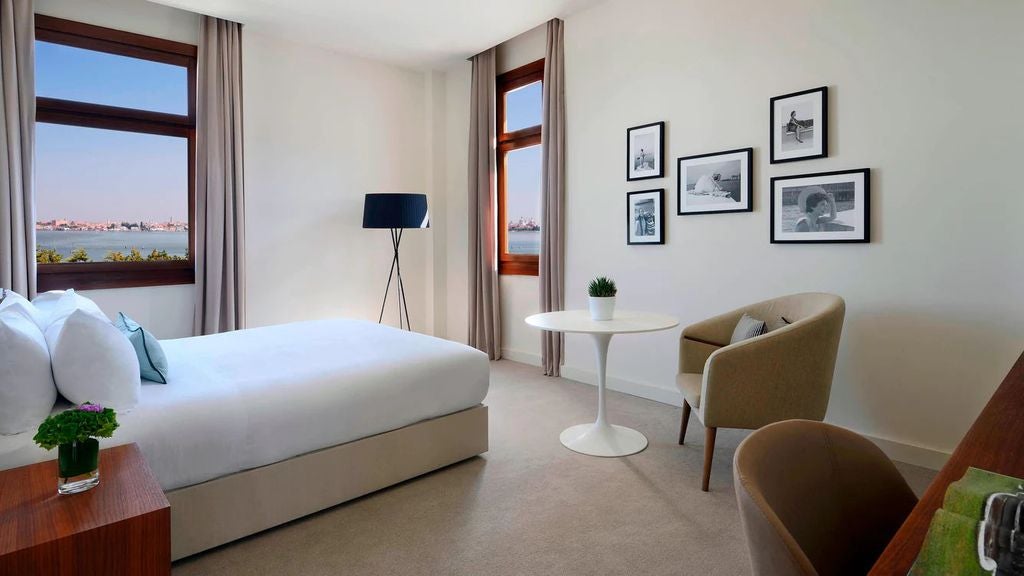 Elegant hotel room with king bed, crisp white linens, floor-to-ceiling windows overlooking Venice lagoon, and modern designer furnishings