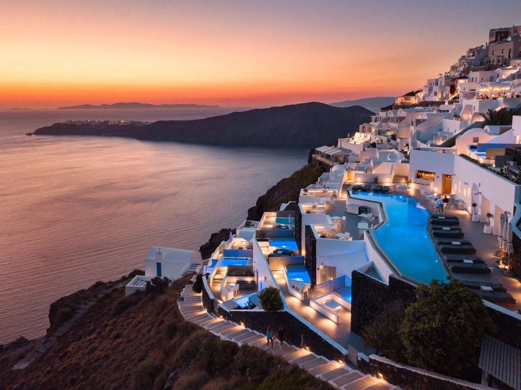 Elegant white hotel perched on Santorini cliffs, featuring infinity pools and terraced balconies overlooking the Aegean Sea at sunset