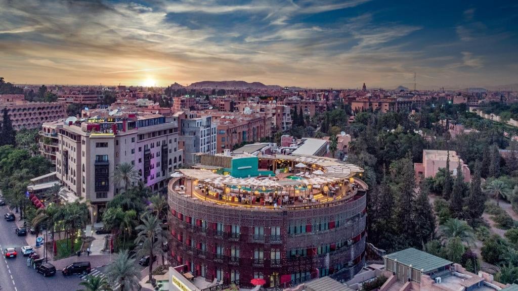 Elegant white-stone Moroccan hotel with arched windows, rooftop terrace, and palm trees set against Atlas Mountains at golden hour