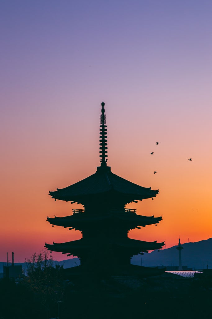 Traditional Japanese pagoda with multiple red-tiled roofs rising above autumn maple trees at golden hour in Kyoto's historic district