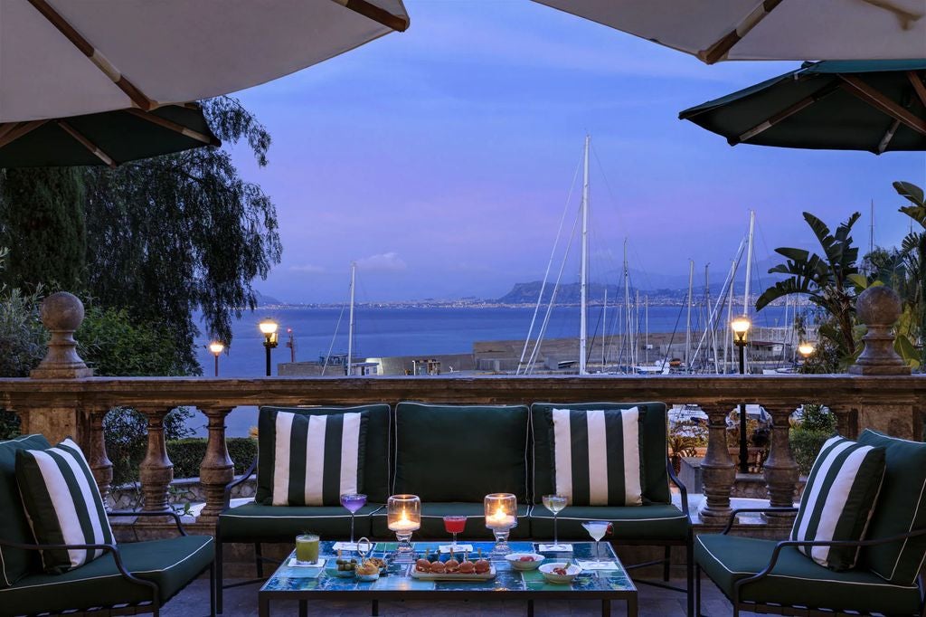 Elegant 19th century Mediterranean palace hotel with grand terrace overlooking Palermo's coastline at sunset, framed by palm trees