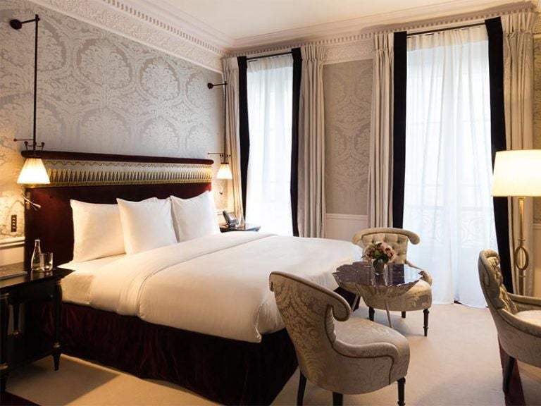 Elegant Parisian hotel room with gold accents, crystal chandelier, antique furniture, draped windows and plush king bed in cream tones