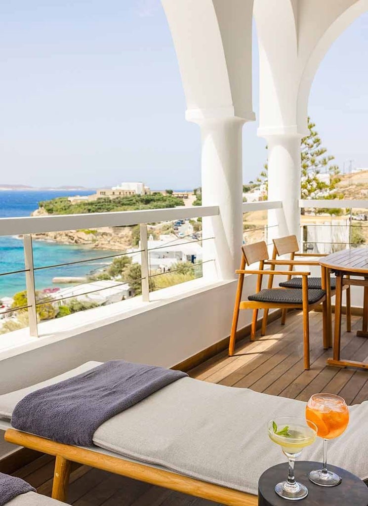 Luxurious white-themed premium suite with panoramic Aegean Sea view, elegant minimalist decor, king bed, and private balcony overlooking Mykonos coastline