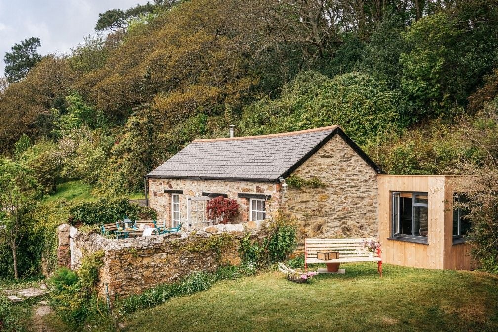 Historic English cottage with ivy-covered stone walls, leaded bay windows, and manicured garden featuring blooming rose bushes and topiaries