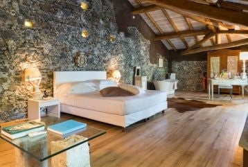 Elegant Sicilian estate suite with rustic stone walls, minimalist design, king bed, and panoramic view of Mount Etna's volcanic landscape at twilight.