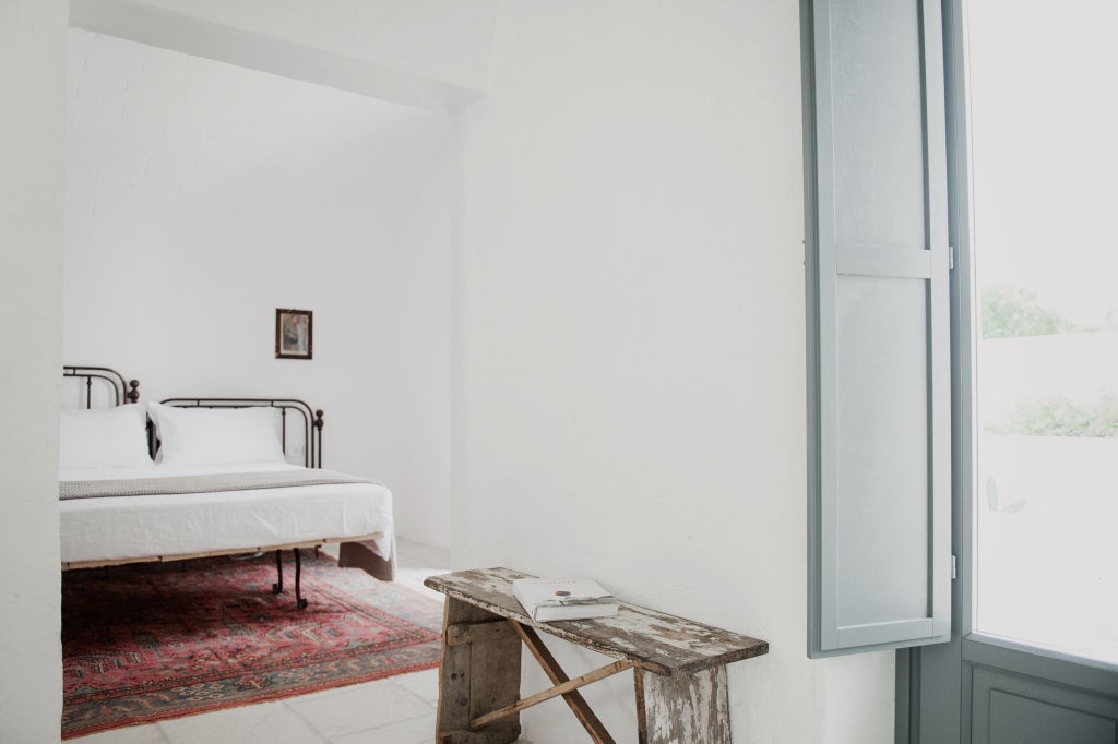 Minimalist white-washed stone room with arched windows, natural linen bedding, and rustic wooden furniture overlooking Italian countryside landscape