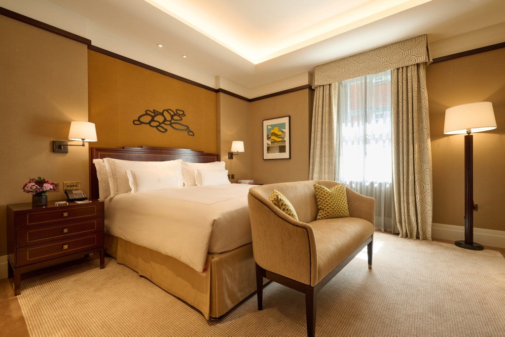 Elegant Mayfair hotel suite with plush king bed, contemporary gold-accented furnishings, soft neutral tones, and panoramic city view windows