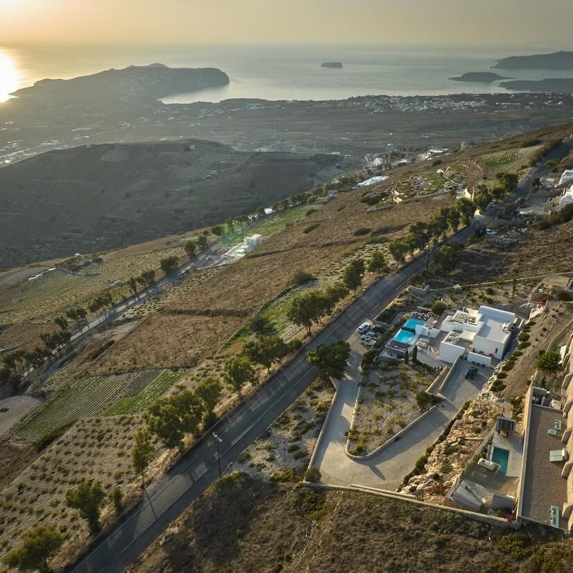 White-washed luxury villa with blue-domed roof overlooking azure Aegean Sea, cascading bougainvillea, and dramatic Santorini cliffside landscape at sunset