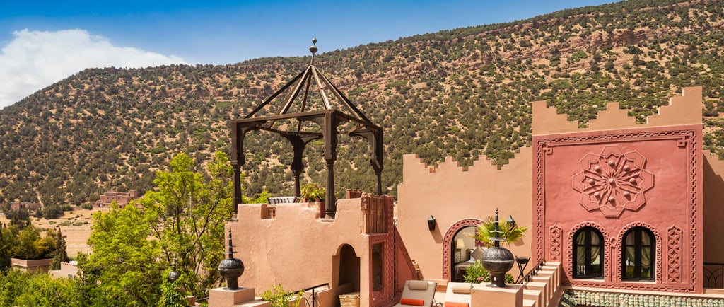 Luxurious Moroccan hotel nestled in Atlas Mountains with traditional red stone architecture, lush gardens and mountain backdrop