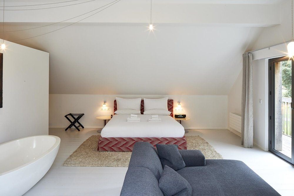 Luxurious loft suite with exposed wooden beams, modern minimalist design, plush white bedding, and panoramic windows overlooking serene countryside landscape